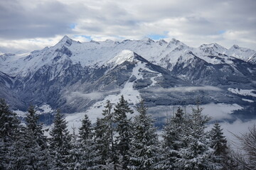 Fototapeta premium Winter landscape with snow-covered trees and mountainsSnow-covered mountains and coniferous forests create a beautiful winter landscape. The cloudy sky and light fog enhance the atmosphere of winter