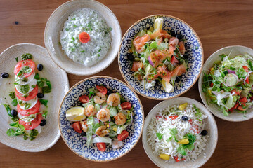 A variety of fresh salads including caprese shrimp and smoked fish are arranged on a wooden table