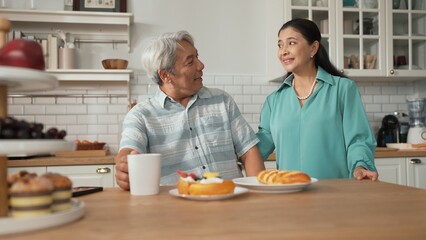Smiling grandfather with casual cloth sitting at table while grandmother presenting breakfast. Senior couple talking and spending time or supporting together. Healthy relationship concept. Myrmidon.