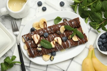Delicious chocolate crepes with banana and blueberries served on white marble table, flat lay