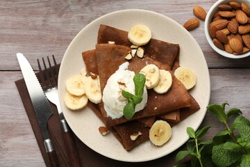 Delicious chocolate crepes with banana, nuts, mint and scoop of ice cream on wooden table, flat lay