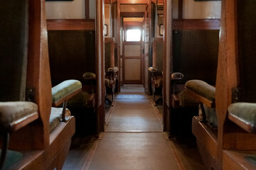 Atmospheric interior of vintage train carriage with wooden panels and green upholstered seats, symmetrical rows leading to bright doorway. Warm lighting, nostalgic ambiance of empty compartment.
