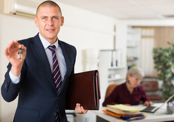 Portrait of cheerful man in formal suit standing in office, holding briefcase with papers and showing key. Successful real estate business concept