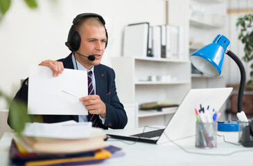 Focused man with headset communicating with clients using video conference application at office