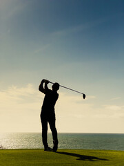 Silhouette of a male golfer mid-swing on a scenic coastal course, bathed in warm golden light. The...