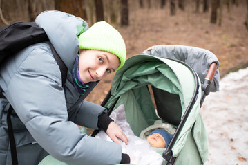 Teenage boy covers his younger brother with a blanket in a stroller. Older brother in a jacket walks with a stroller with a baby in a park in spring or winter.