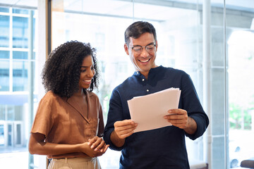 Business team of two happy young busy diverse colleagues, professional business man and business woman executives working together standing in office looking at project documents and talking.