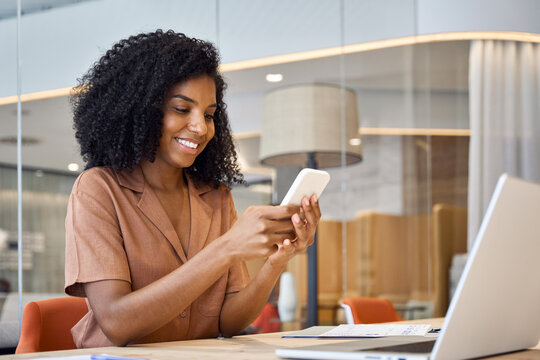 Happy young African American business woman holding smartphone sitting in office, smiling ethnic businesswoman using financial banking app on cell phone looking at cellphone working with mobile tech.