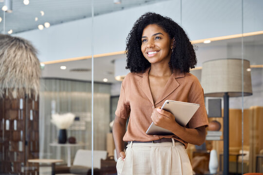 Happy confident young African American professional woman, female entrepreneur, businesswoman looking away holding tab standing in office at work dreaming of future success.