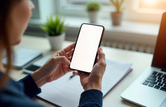 Woman holds blank screen smartphone in office. Modern workplace. Businesswoman uses mobile phone. Focus on device. Hands on phone. White table. Laptop visible. Closeup view. Business tech. Digital