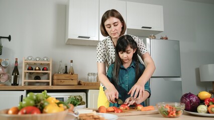 Smart caucasian mother and asian girl cooking together and chopping vegetable or preparing salad for dinner. Happy mom and daughter making healthy food with fresh food. Healthy food concept. Pedagogy.