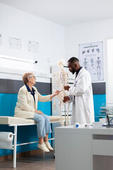 Obraz premium Orthopedic specialist shows skeleton model to retired female patient discussing human anatomy and medical options. African american male doctor describes treatment process to senior white woman.