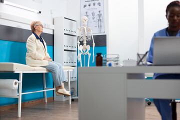 Selective focus of caucasian senior woman patiently waiting on examination bed for health checkup. Elderly lady seated in hospital room ready for her medical appointment, as black nurse works nearby.