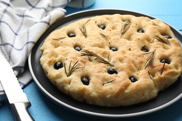 Fototapeta premium Delicious focaccia bread with olives, rosemary and knife on light blue wooden table, closeup