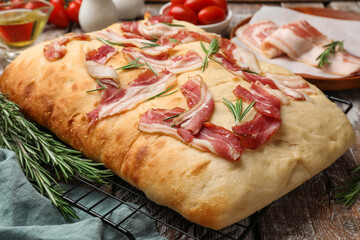 Delicious focaccia bread with bacon, rosemary, oil and tomatoes on wooden table, closeup