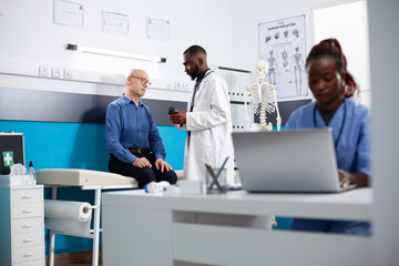 Friendly physician holds prescription bottle while explaining treatment plan to elderly male patient. Caucasian retired man sits in clinic office receiving medication from doctor for his recovery.