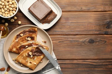 Tasty crepes with chocolate butter, nuts, honey and knife on wooden table, flat lay. Space for text
