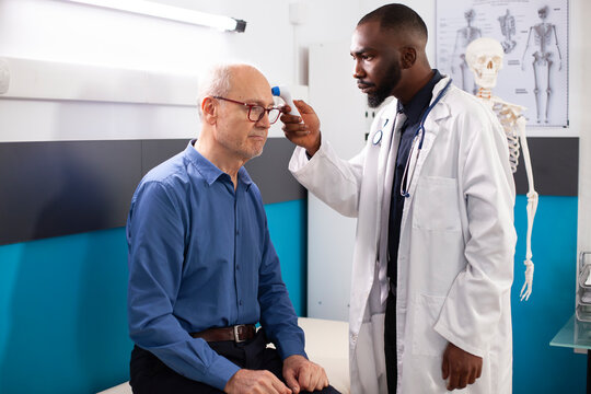 Professional male physician uses thermometer on elderly patient in hospital room. Caucasian old man seated while african american doctor checks his temperature during routine medical checkup.