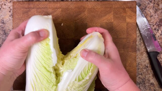 The napa cabbage is cut in half lengthwise with a knife and finely chopped for the salad on a wooden cutting board. Top view.