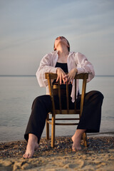 Young woman in relaxed pose, sitting on chair by calm beach, embodying serenity. Tranquil seascape...