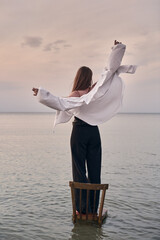 Young woman joyfully stretches her arms, standing on wooden chair in calm waters. Scene evokes...
