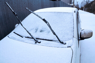 The windshield of the car with the wipers turned up, covered with fresh snow. Car in the snow close-up on the background of a country houses, winter day.