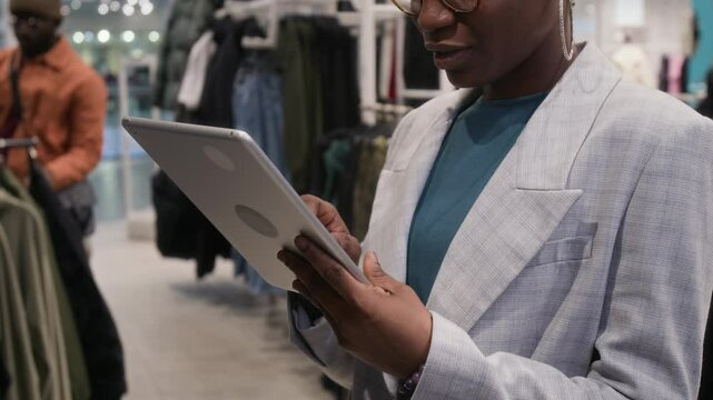 Side cropped shot of young Black female fashion stylist in checkered blazer, glasses and with big ring earrings using tablet computer while working in clothes shop