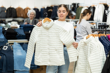 Pleased young girl choosing white jacket in clothing store with large assortment