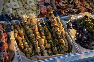 Variety of grilled food on display at market or food stall. There are skewers of chicken, vegetables, and seafood, as well as grilled mushrooms
