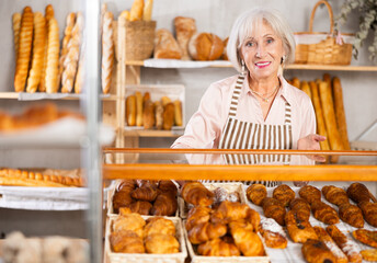 Elderly bakery saleswoman places fresh hot croissants on display