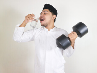 Excited Asian Muslim man holding a dumbbell and drinking a glass of water. concept of a healthy lifestyle for Islamic religious people in the context of the month of Ramadan and Eid Mubarak