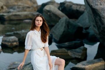 Serene woman in white dress sitting on rocky shore, gazing at tranquil ocean view
