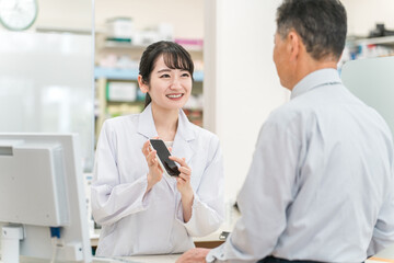 Fototapeta premium 薬局で高齢者・中高年男性にスマホアプリを説明する薬剤師・調剤師の女性（服薬指導） 