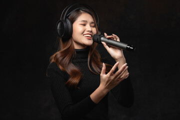 Obraz premium Beautiful female singer with long brown hair with headphones singing karaoke with microphone on stage in dark background.