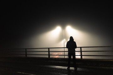 A sinister hooded figure, standing above a motorway. On a mysterious foggy night. Silhouetted by street lights