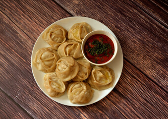 Steamed manti on a large round ceramic plate with a cup of spicy tomato sauce on a wooden table.