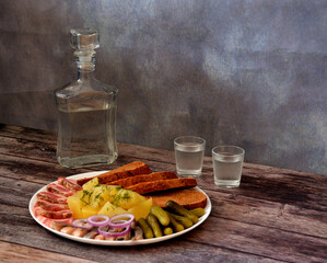A glass decanter and two glasses of cold vodka on a wooden table, next to a plate with potatoes, lard, cucumbers, herring and rye bread.