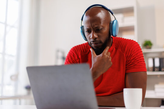 Young beautiful African American man in headphones working with laptop at office at workplace, joyful and satisfied business man watching online webinar, educational video course, learning remotely.