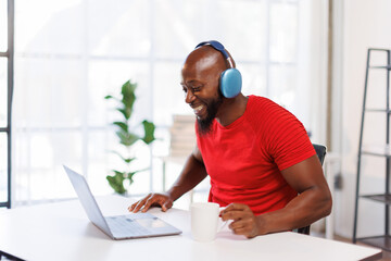 Young beautiful African American man in headphones working with laptop at office at workplace,...