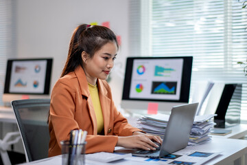 A woman in an orange jacket is working on a laptop in front of a monitor