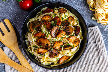 A delicious serving of creamy tagliatelle pasta topped with golden chicken and sautéed mushrooms, garnished with fresh parsley. Presented in a black bowl on a rustic background.