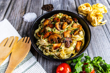 A delicious serving of creamy tagliatelle pasta topped with golden chicken and sautéed mushrooms, garnished with fresh parsley. Presented in a black bowl on a rustic background.