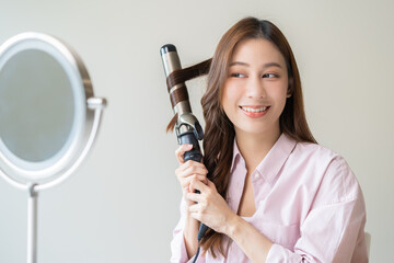 Beautiful curly brunette, attractive asian young woman, girl smiling using curler, curling iron long hair with ceramic curler straightener at home. Hairstyle and Hairdressing. Beauty and hair care.