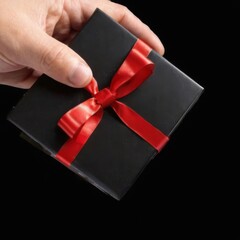 Black gift box with red ribbon, held elegantly in hand on black background, shiny material, perfect for gifting.
