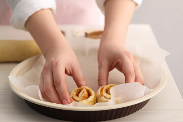 Woman putting cinnamon roll into baking dish at white wooden table, closeup