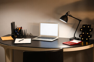 Blank laptop with stationery and glowing lamp on table in office at night