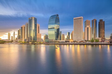Fototapeta premium A modern city skyline with tall glass skyscrapers reflecting in the calm waters of a river or bay at sunset