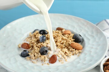 Pouring milk into bowl with oatmeal, blueberries and almonds at light blue table, closeup