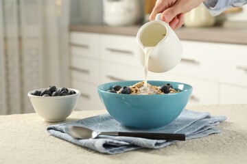 Woman pouring milk into bowl with oatmeal and blueberries at beige textured table, closeup