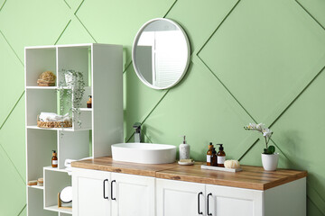 Stylish interior of bathroom with shelving unit, chest of drawers and sink near green wall